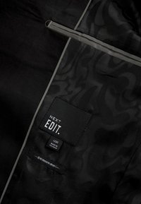 Černý vnitřek oděvu se slabým tonálním vzorem, obsahující štítky: "NEXT EDIT," velikost "40R" a "SKINNY FIT." Ozdoben šedými akcenty.