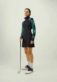 Moteris juodai ir žaliai ilgomis rankovėmis suknele laiko golfo lazdą, vilkėdama baltas kojines ir juodai baltus golfo batus, stovinti ant paprasto fono.