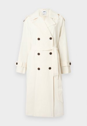 Crèmekleurige double-breasted trenchcoat met donkere knopen, tailleceintuur, schouderepauletten en manchetbandjes tegen een lichte achtergrond.