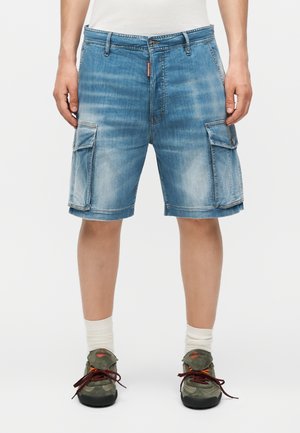 Denim cargoshorts med sidelommer båret med hvide sokker og grønne snørebåndssneakers på en person, der står.