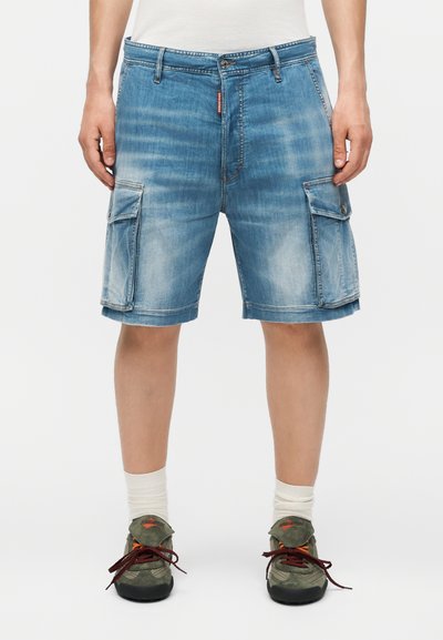Short cargo en denim avec poches latérales à rabat, porté avec des chaussettes blanches et des baskets vertes à lacets sur une personne debout.