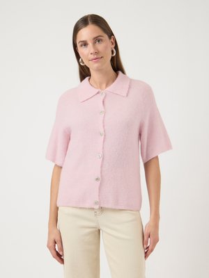 Femme portant un pull à boutons en maille duveteuse rose clair à manches courtes et un pantalon blanc cassé, debout devant un fond clair uni.