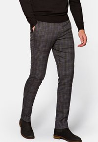 Mężczyzna ubrany w brązowe, wąskie spodnie w kratę z niebieskimi liniami, czarny sweter i czarne zamszowe botki na drewnianej podeszwie, ręce swobodnie opuszczone.