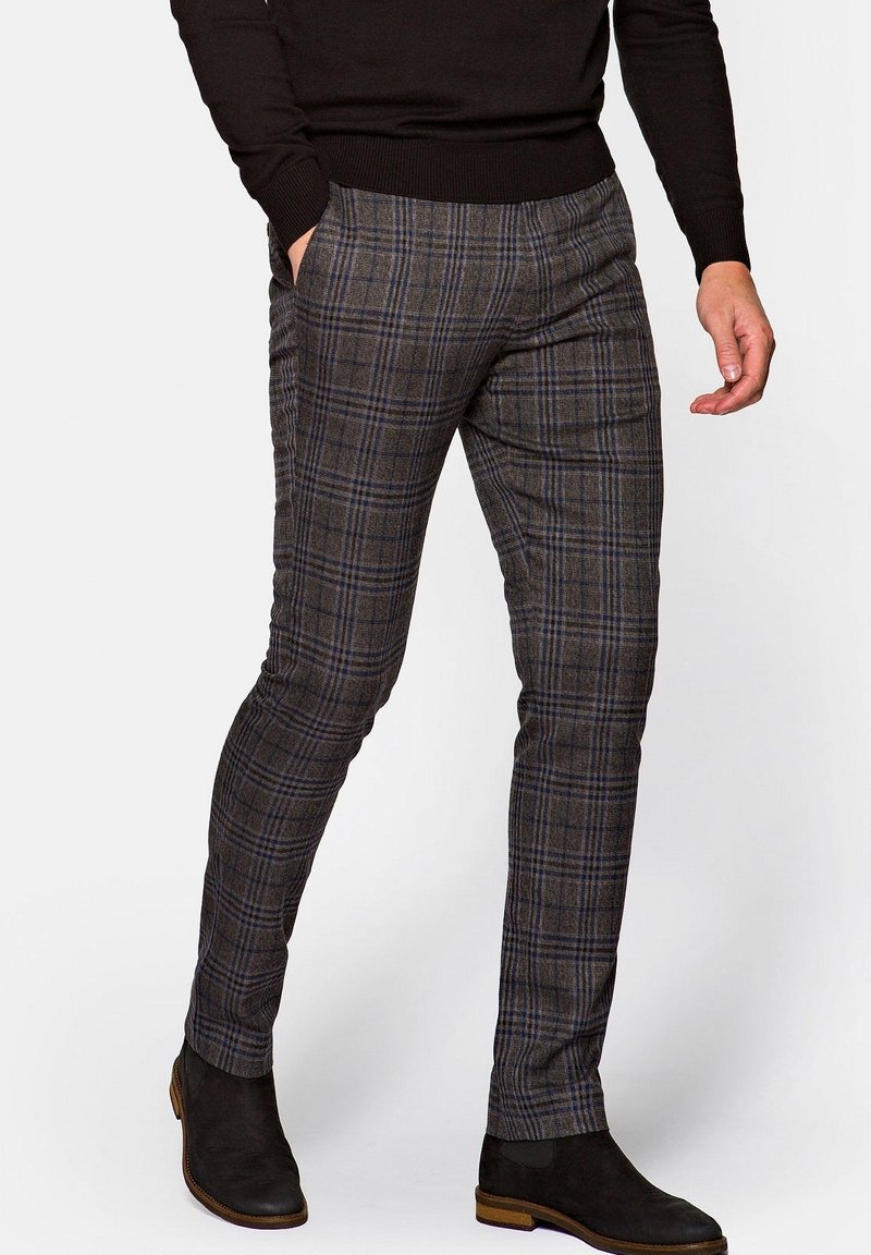 Mężczyzna ubrany w brązowe, wąskie spodnie w kratę z niebieskimi liniami, czarny sweter i czarne zamszowe botki na drewnianej podeszwie, ręce swobodnie opuszczone.