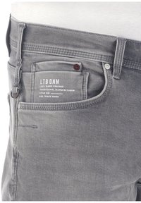 Jeans de mezclilla gris con diseño de cinco bolsillos, costuras visibles, un botón decorativo en forma de círculo y un bolsillo trasero etiquetado con la marca.