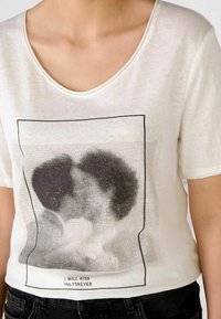 Hvid bomulds T-shirt med korte ærmer, rund halsudskæring, der har et sort prikket grafisk print af to ansigtter, der kysser, samt tekst nedenfor.