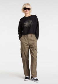 Zwarte cropped longsleeve met een gouden spiraal logo, gecombineerd met olijfkleurige cargobroeken met zijzakken en zwarte Vans sneakers.