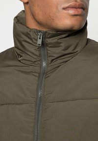 Puffjacka i mörkgrön tyg med hög krage, utrustad med mittdragkedja och texturerad yta. Inga ytterligare mönster synliga.