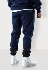 Marineblaue Sweatpants mit elastischen Bündchen, ausgestattet mit Seitennaht und einem kleinen gestickten Logo auf der Rückseite. Kombiniert mit weißen Sneakers.