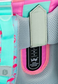 Detail eines Rucksacks mit einem türkisfarbenen Gurt, grauem Netzpanel, rosa Akzenten und einem schwarzen Etikett, das "baagl Ergo Adjustable System" mit Größenmarkierungen anzeigt.
