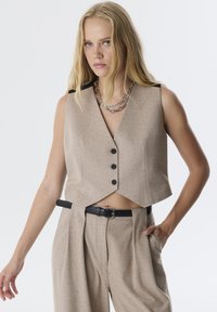 Beige cropped vest met een V-hals, zwarte schouderaccenten en vijf knopen. Gecombineerd met losse broek en een zwarte riem.