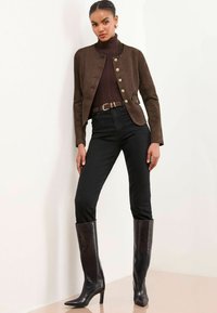 Veste en daim marron avec boutons dorés, col roulé noir ajusté, jeans noirs et bottes en cuir noires jusqu'aux genoux. Fond uni.