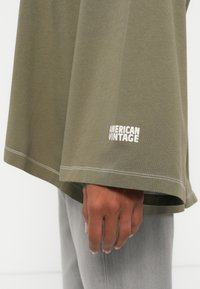 Ελιά πράσινο υπερμεγέθες πουκάμισο από βαμβάκι με λευκή κεντητή επιγραφή "AMERICAN VINTAGE", διαθέτοντας φαρδιές μανσέτες και άνετη εφαρμογή.