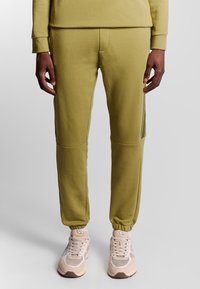 Olivgrüne Jogginghosen mit Seitenelementen, elastischen Bündchen und schmaler Passform, kombiniert mit hellbeigen Sportschuhen.