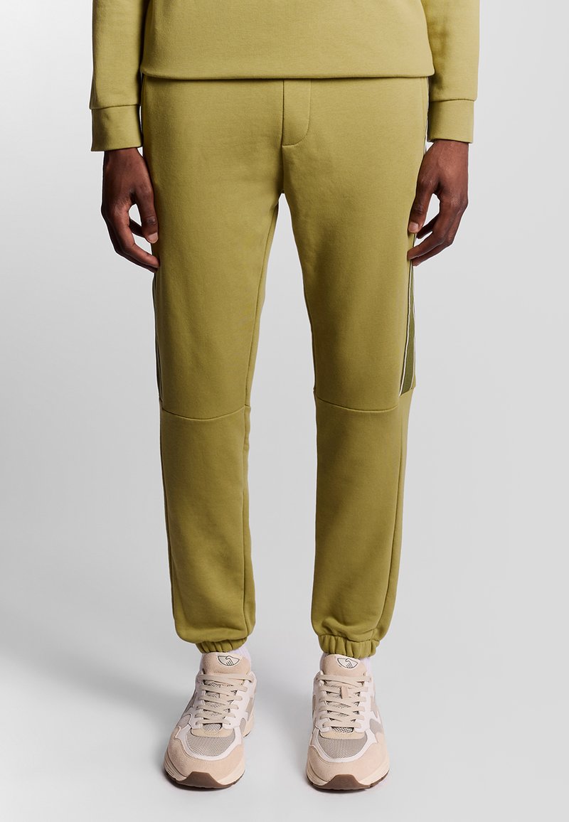 Olivgrüne Jogginghosen mit Seitenelementen, elastischen Bündchen und schmaler Passform, kombiniert mit hellbeigen Sportschuhen.