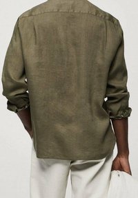 Olijfgroene linnen blouse met lange mouwen, met knoopmanchetten en een rugpatroon, gecombineerd met lichtgekleurde broeken, vastgehouden met één hand.