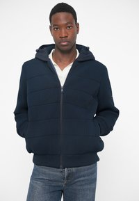 Ναυτικό μπλε φούτερ με φερμουάρ και ριχτό σχέδιο, ρυθμιζόμενη κουκούλα και εμπρός τσέπες. Φορεμένο πάνω από ένα λευκό πουκάμισο και σε συνδυασμό με μπλε τζιν.