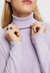 Hellblauer Rollkragenpullover mit geripptem Muster, taillierter Passform und langen Ärmeln. Hände, die den Kragen anpassen und mehrere Ringe tragen.