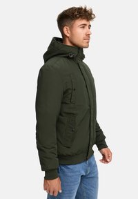 Chaqueta capucha verde oscuro con cierre de cremallera al frente, puños acanalados y múltiples detalles de bolsillo, combinada con jeans azul claro.