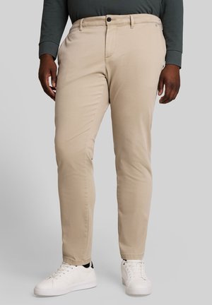 Personne portant un pantalon beige, une chemise à manches longues vert foncé et des baskets blanches, debout devant un fond clair uni.