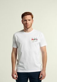 Camiseta de algodón blanca con un diseño gráfico a color que incluye texto y banderas en el área del pecho izquierdo. Cuello redondo y mangas cortas.
