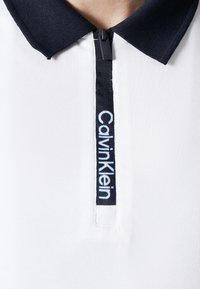 Bílá polokošile s námořnickým límcem, se zipovým zapínáním a štítkem s nápisem "Calvin Klein" modrými písmeny a texturovanou tkaninou.