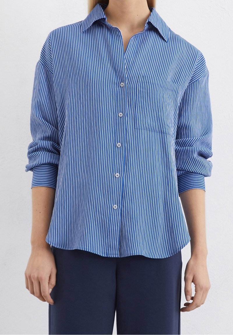 Femme portant une chemise à rayures verticales bleues et blanches, manches retroussées, pantalon foncé, debout contre un mur uni.