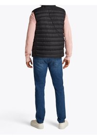 Czarna pikowana kamizelka na jasnoróżowej koszuli z długim rękawem, w połączeniu z niebieskimi dżinsami. Widoczne białe sneakersy; prosty i dopasowany design.