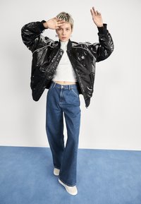Giacca bomber nera lucida con strisce bianche, top bianco corto e jeans a gamba larga blu. La modella indossa scarpe da ginnastica di colore chiaro.