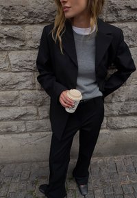 Melnā pārāk liela bleizeri virs pelēka džempera, apvienojot ar melnu biksēm. Modelis tur kafijas krūzi un valkā melnus augstpapēžu klumpes.