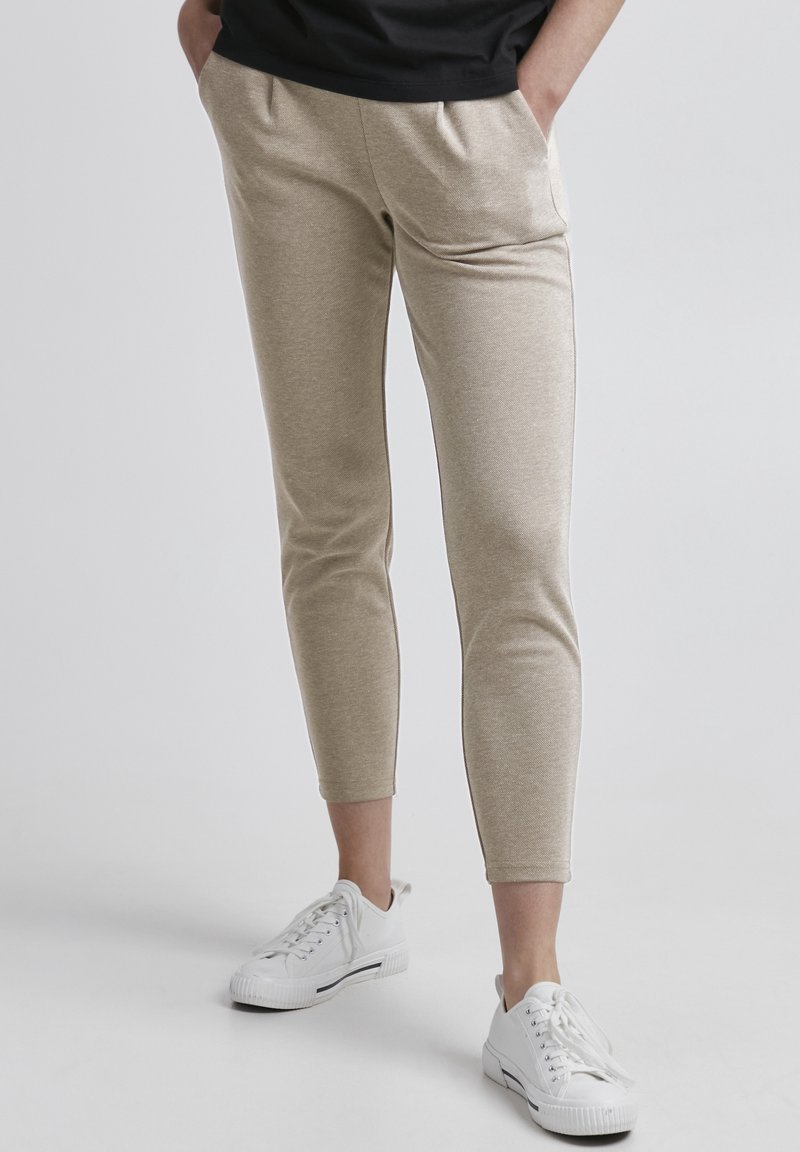 Pantalons de survêtement beige, fuselés, en tissu doux, avec une taille élastique et des poches latérales, associés à des chaussures de sport blanches.