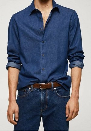 Mand iført en mørkeblå denimskjorte med opkrogne ærmer, stoppet ind i blå jeans med et brunt læderbælte.