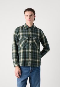 Green and white plaid flannel shirt with long sleeves, button-down collar, two chest pockets, and textured fabric. Casual fit.