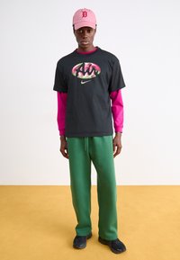 Svart grafisk T-shirt med "Air"-logotyp, lager på lager med en rosa långärmad tröja, kombinerad med gröna träningsbyxor och svarta sneakers. Rosa keps ovanpå.