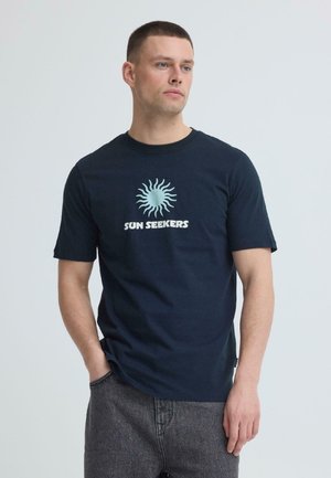 Mies pukeutuneena tummansiniseen "SUN SEEKERS" t-paitaan, jossa vaaleansininen aurinkografiikka, seisoo oikea käsi harmaiden farkkujen taskussa ja katsoo oikealle.