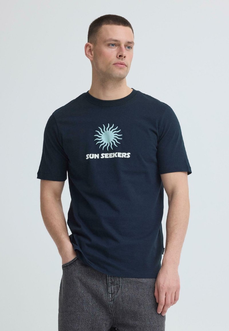 Άνδρας που φοράει μπλε μπλουζάκι "SUN SEEKERS" με γαλάζιο γραφικό ήλιο, στέκεται με το ένα χέρι στην τσέπη του γκρι τζιν, κοιτάζοντας δεξιά.