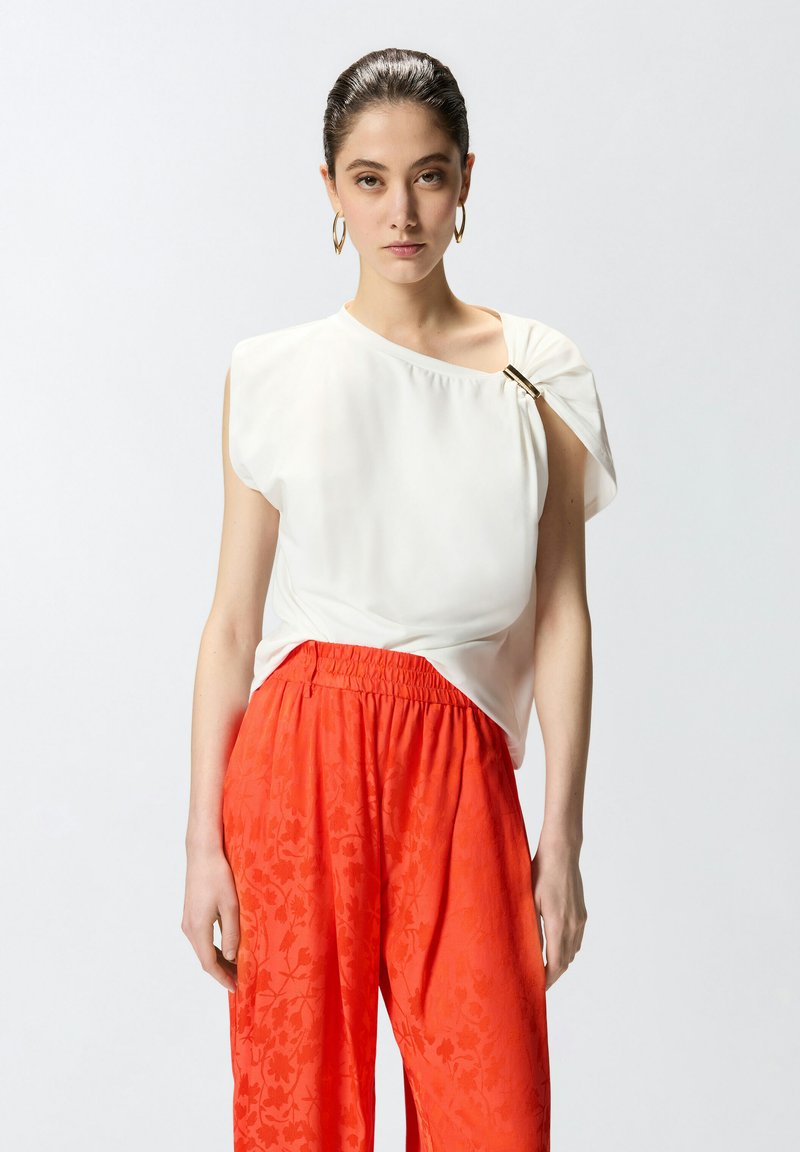 Ženska z zglajenimi lasmi, ki nosi zlate obroče, belo asimetrično majico s sponko in rdeče cvetlične hlače visokega pasu in ohlapnega kroja.