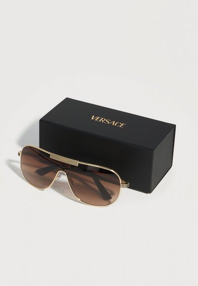 Lunettes de soleil aviateur à monture dorée avec verres bruns, posées à côté d'une boîte noire Versace avec lettrage doré, sur une surface blanche.