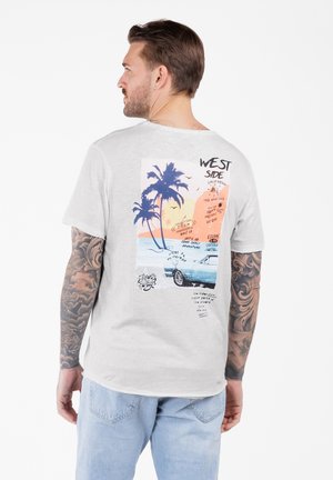 Mann mit Tattoos an beiden Armen, der ein weißes T-Shirt trägt, das auf der Rückseite einen bunten Grafikdruck mit einem Palmenbaum, einem Auto und Text zeigt.