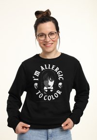 Donna con occhiali e capelli raccolti in un chignon, che indossa una felpa nera con la scritta "Sono allergica al colore" accompagnata da un volto femminile in stile gotico e teschi.