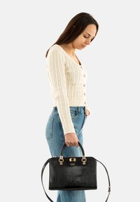 Sac à main noir avec une texture crocodile, une quincaillerie en or et une bandoulière détachable, tenu par une personne portant un pull côtelé crème et un jean bleu.