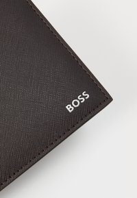 Portafoglio di un marrone scuro con texture e un motivo sottile. Logo "BOSS" bianco nell'angolo in basso a destra, che contrasta con il materiale e le cuciture.