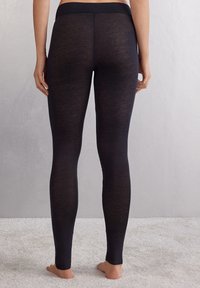Svarta leggings med hög midja, slät textur och åtsittande design, visade från baksidan, mot en mjuk, ljus yta.