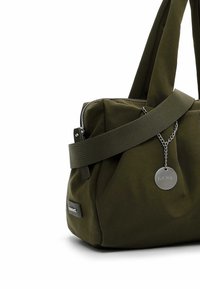 Sac en tissu doux vert foncé avec une large bandoulière, une fermeture éclair sur le dessus, une étiquette ronde en métal sur chaîne et une petite étiquette rectangulaire.