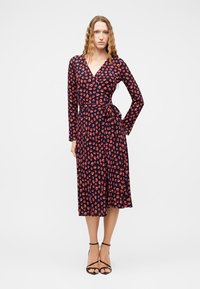 Γυναίκα με μακριά κυματιστά μαλλιά, ντυμένη με μεσάτο navy φόρεμα τύπου wrap με κόκκινο λουλουδάτο σχέδιο και μαύρες ψηλοτάκουνες πέδιλα, στέκεται με το ένα χέρι στη μέση.