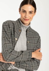 Veste à carreaux grise avec des boutons dorés, texture douce, et un poignet froncé, portée sur un pull à col roulé gris.