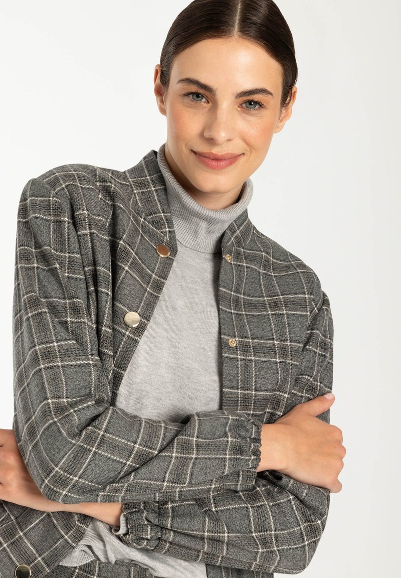 Veste à carreaux grise avec des boutons dorés, texture douce, et un poignet froncé, portée sur un pull à col roulé gris.