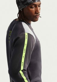 Άνδρας που φοράει σκούρο γκρι φούτερ Nike με λωρίδα λογότυπου νέον κίτρινου στο μανίκι και γκρι καμουφλάζ σκουφί, κοιτώντας προς τα κάτω.