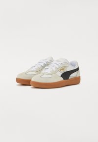 Sneakers bianchi e neri in materiale scamosciato, suola in gomma color tan, lacci bianchi e un logo dorato sul lato. Design sportivo classico.