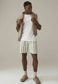 Tenue décontractée comprenant un t-shirt blanc, un sweat-shirt beige porté sur les épaules, un short rayé de couleur claire et des chaussures à enfiler couleur miel.